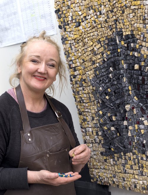 Headshot of Louise in her studio standing in front of her artwork made up of gold and black pieces of mosaic.