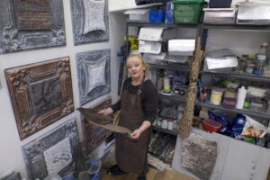 Louise in her studio showing. Holding one of her artworks and surrounded by two walls of sections of her mosaic assemblage artworks and shelves with her construction material she uses to make the work.