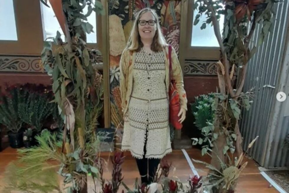 A picture of Angela at a flower show wearing earthy coloured clothes around a decorative border of native flowers.