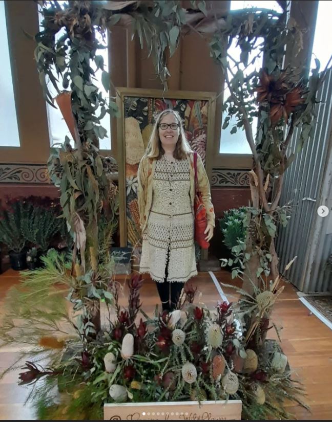 A picture of Angela at a flower show wearing earthy coloured clothes around a decorative border of native flowers.