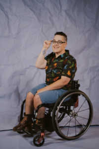 Jax is seated in their manual wheelchair smiling at the camera. They are wearing a black shirt with blue, green, orange and red frogs on it with a black background, blue shorts and maroon boots with rainbow laces. They have short black hair and are wearing glasses. Their right-hand is touching their glasses so you can see their wheelchair feminist tattoo]