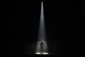 A solo violinist stands on a dark stage, lit only by a dramatic spotlight from above.