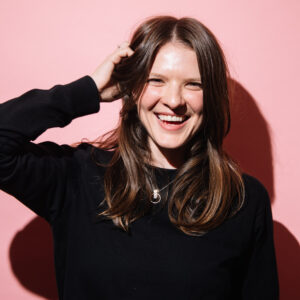 A2 photograph of smiling woman her hand in her brown hair. The photo is on a pink background, it makes me feel excited and happy.