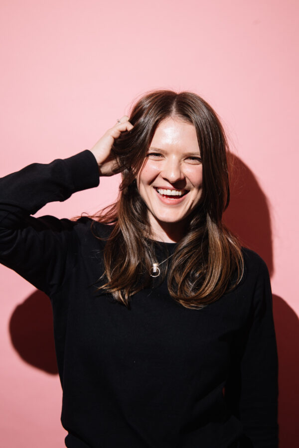 A2 photograph of smiling woman her hand in her brown hair. The photo is on a pink background, it makes me feel excited and happy.