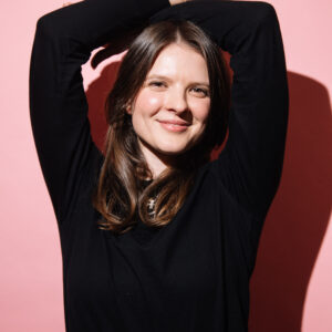 A2 sized photograph of a woman with a pink background, she has her hands on her head. She looks as though she is happy.