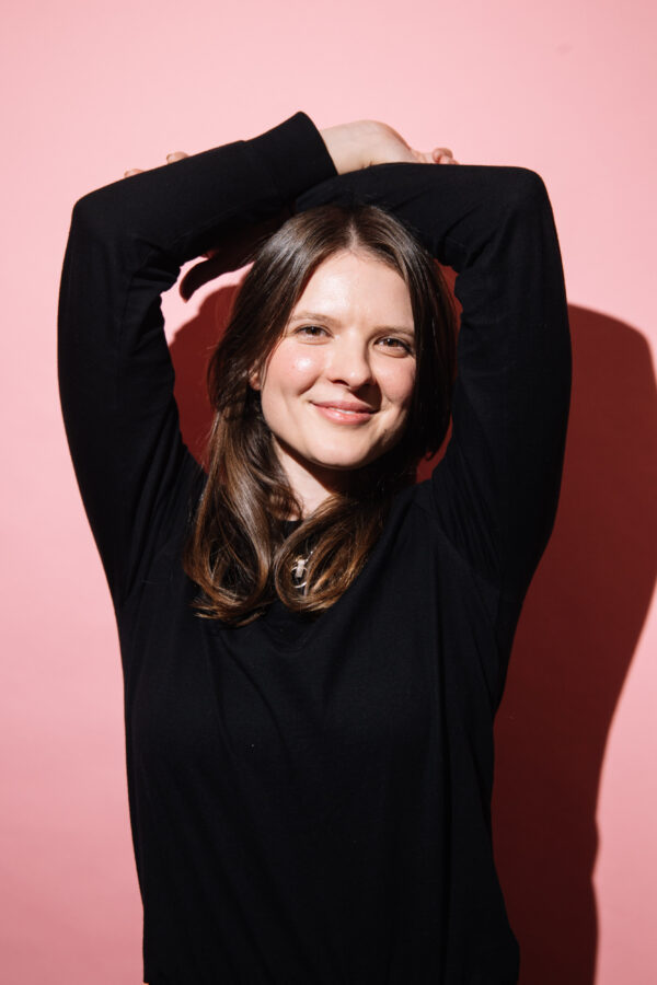 A2 sized photograph of a woman with a pink background, she has her hands on her head. She looks as though she is happy.