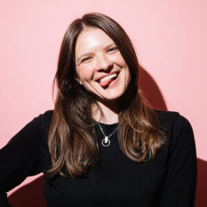 A2 photograph of a woman with a pink background. She has her tongue out and her head is tilted to one side with her hand on her hip, this image makes me feel funny and silly.