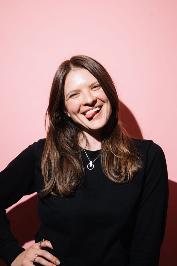 A2 photograph of a woman with a pink background. She has her tongue out and her head is tilted to one side with her hand on her hip, this image makes me feel funny and silly.