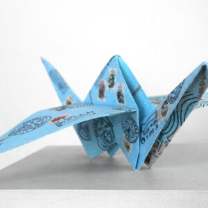 A single folded origami crane. It is a sky blue colour with postage stamps and symbols on it.