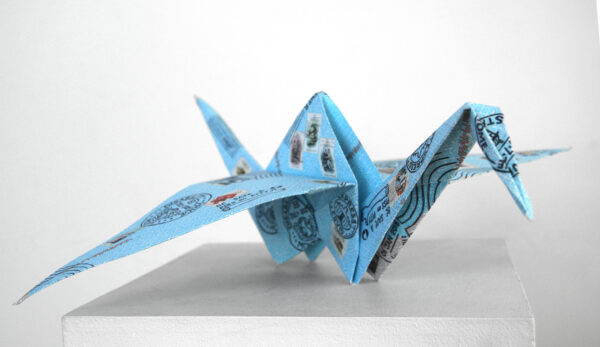A single folded origami crane. It is a sky blue colour with postage stamps and symbols on it.