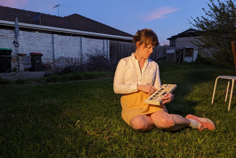 Katie Dey sits on the lawn holding a small keyboard.