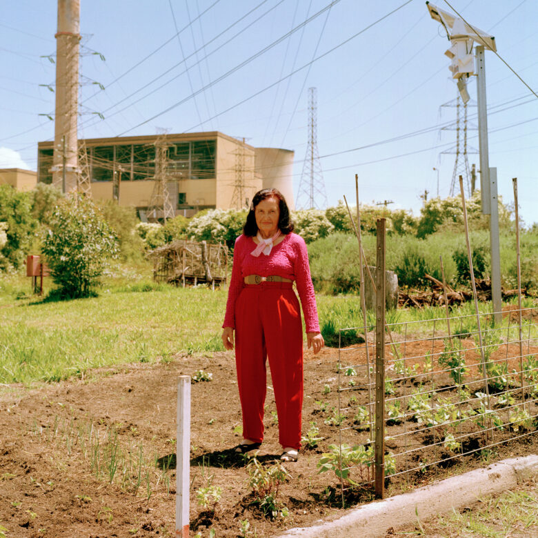 An elderly woman wearing a bright pink long sleeve top and red trousers standing in a patch of land. On her left, there are newly growing plants. There are trees on a green patch of grass behind her, surrounded by power line towers in front of a power station.