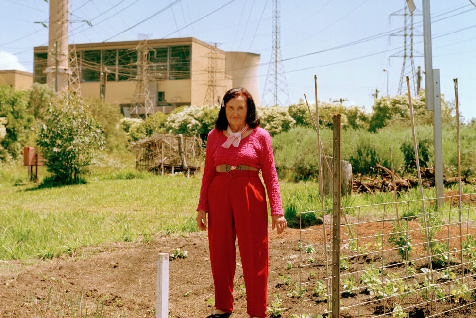 An elderly woman wearing a bright pink long sleeve top and red trousers standing in a patch of land. On her left, there are newly growing plants. There are trees on a green patch of grass behind her, surrounded by power line towers in front of a power station.