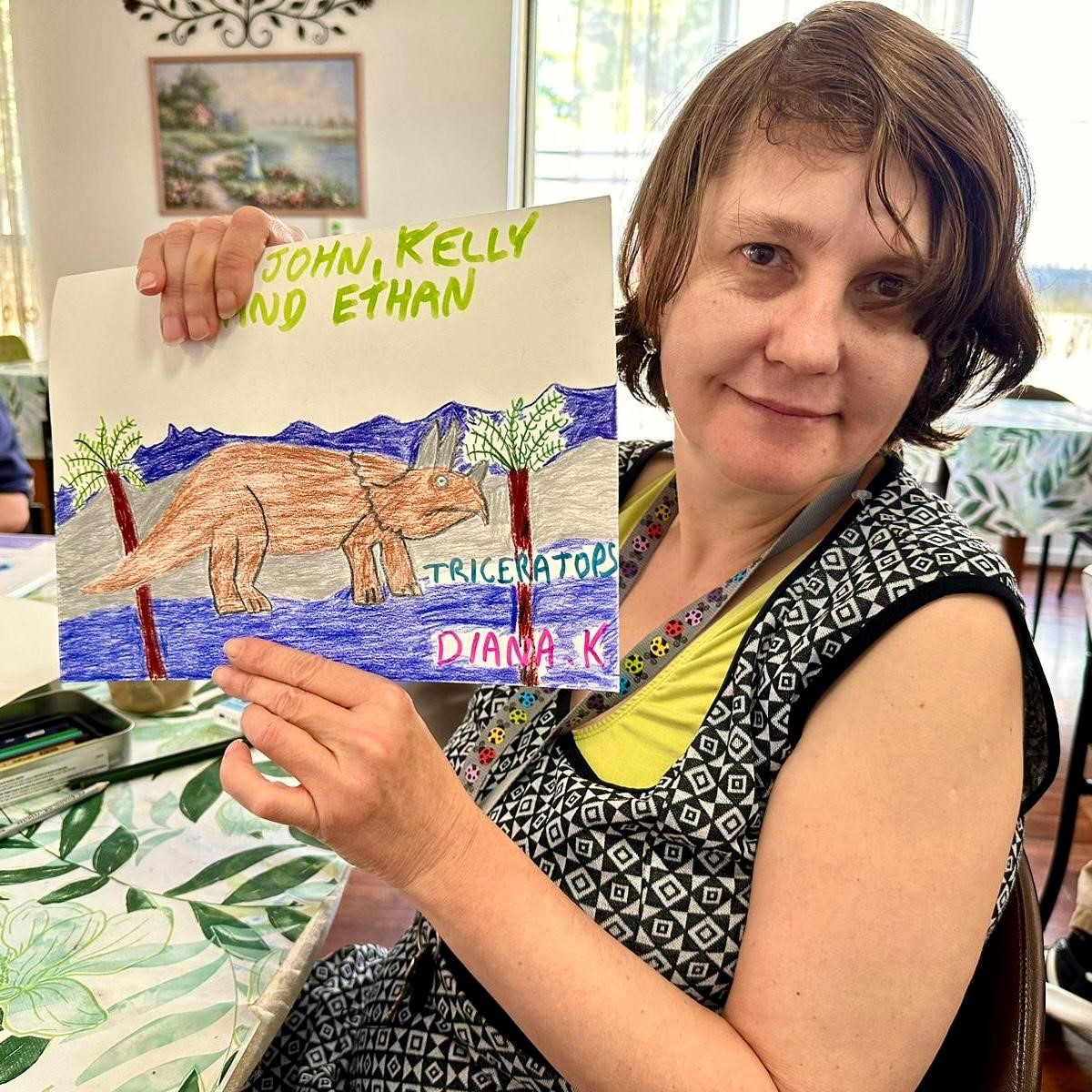 Diana holding her artwork of Triceratops and looking to camera with gentle smile.