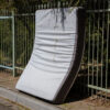 A double mattress on the curb leaning vertically against a green fence