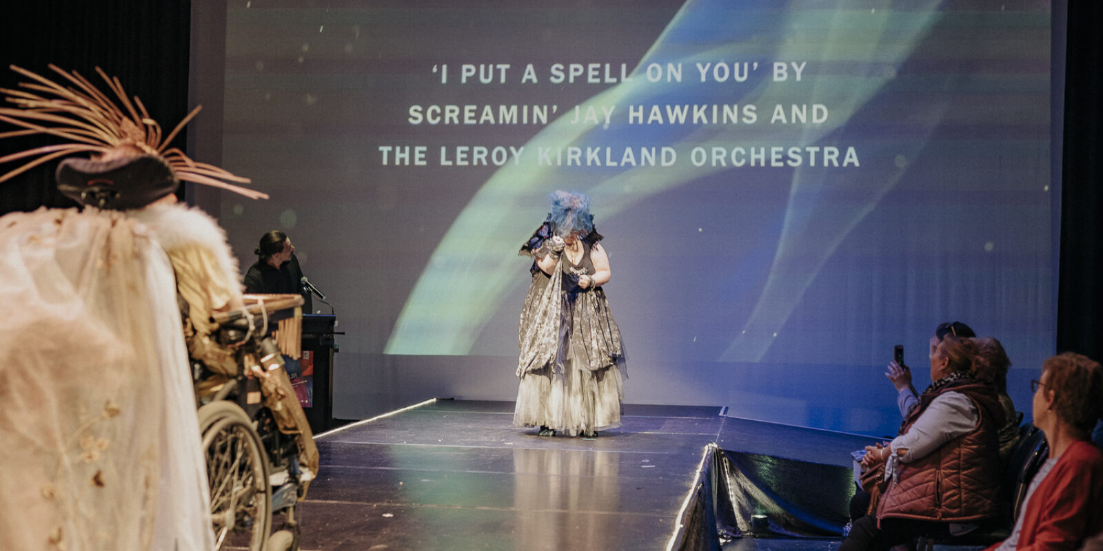 Two people in costumes on a runway stage with audience members seated side of stage. A projection behind them reads 