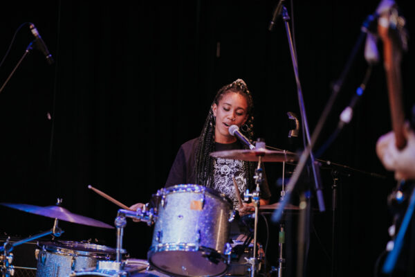 A person performing seated behind drums and microphone on stage.