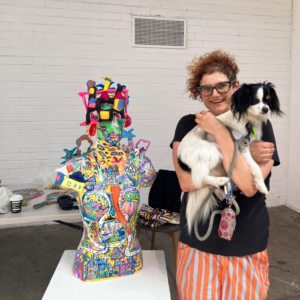 A person holding a small dog next to a manniquin covered in collage paper and colourful materials.