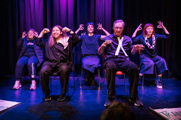 Five people seated on stage gesturing forward.