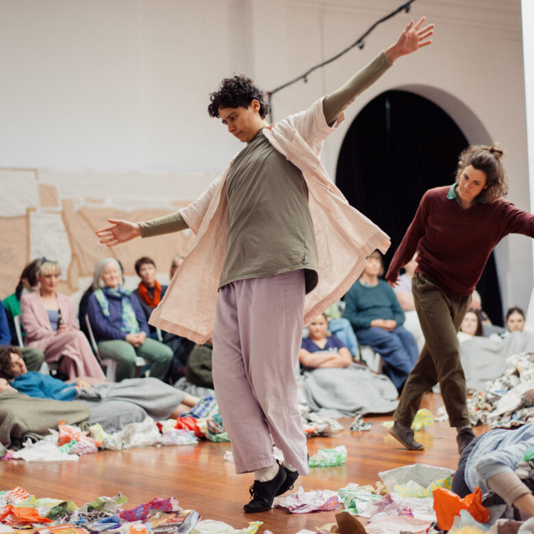 Contemporary dancers perform amid colorful scattered materials in a gallery, while seated spectators watch; the scene feels lively and expressive.
