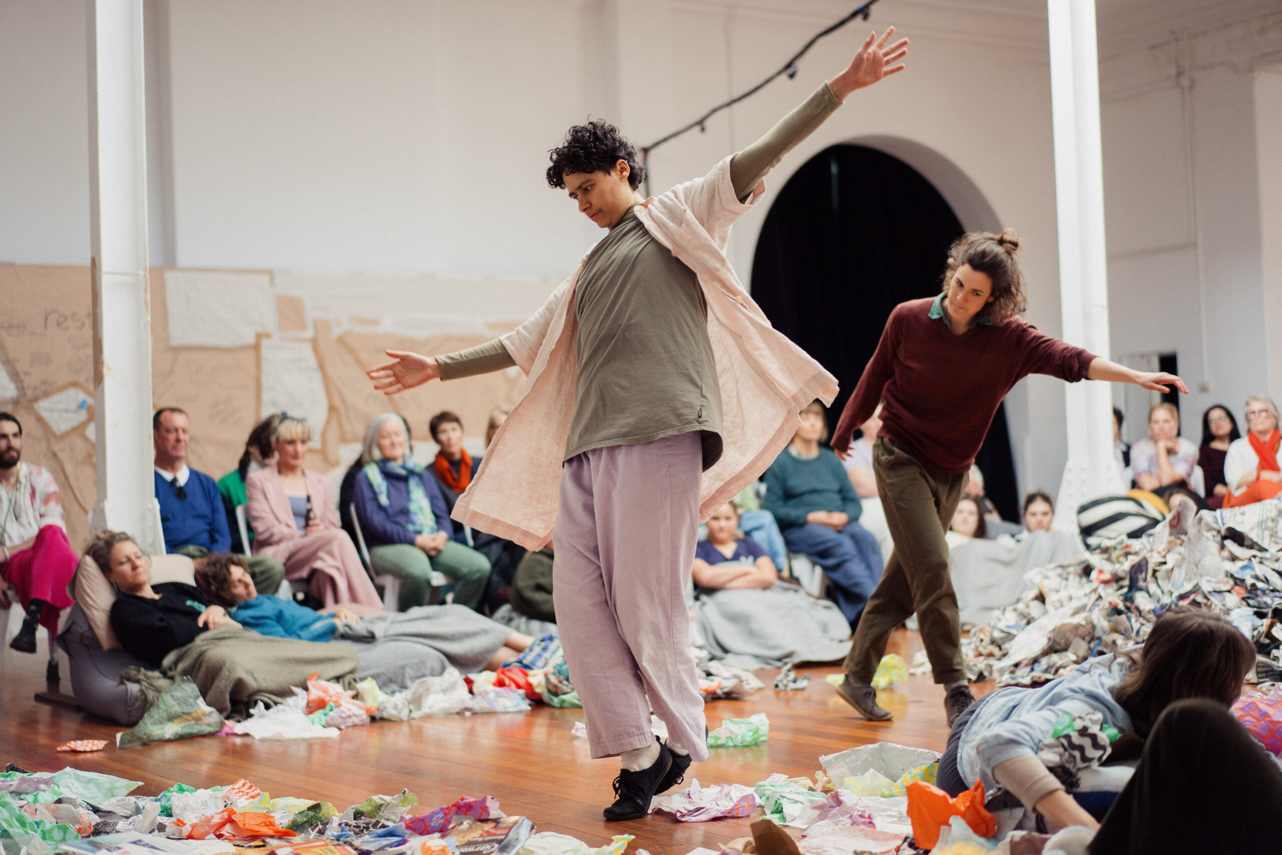 Contemporary dancers perform amid colorful scattered materials in a gallery, while seated spectators watch; the scene feels lively and expressive.