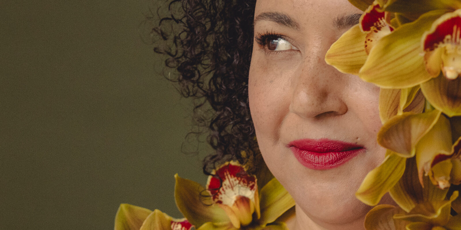 Artist Pearl Blackk (she/her) a brown-skinned woman with dark hair gazes to the right. Her face is hidden behind growing yellow orchids with red centres in full bloom. Behind her is an earthy dark moss background – this image celebrates the concept of regeneration for Alter State 2026.