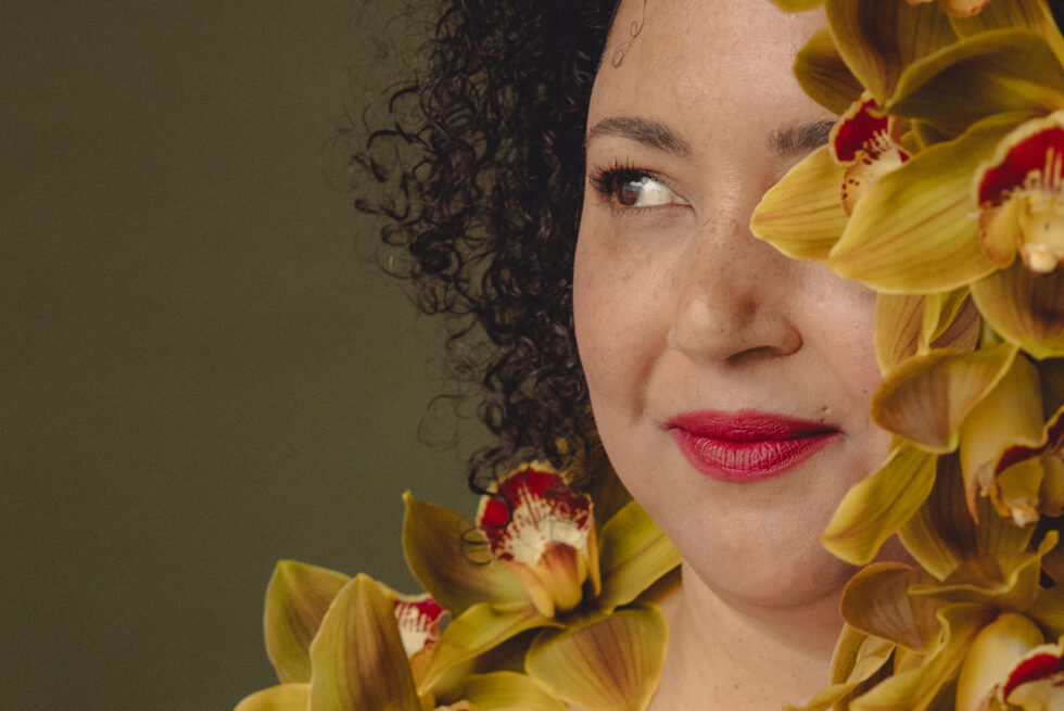 Artist Pearl Blackk (she/her) a brown-skinned woman with dark hair gazes to the right. Her face is hidden behind growing yellow orchids with red centres in full bloom. Behind her is an earthy dark moss background – this image celebrates the concept of regeneration for Alter State 2026.