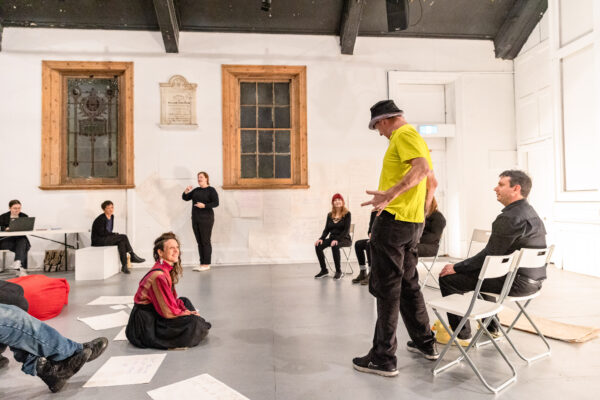 People standing and seated in an open hall rehearsing a performance.
