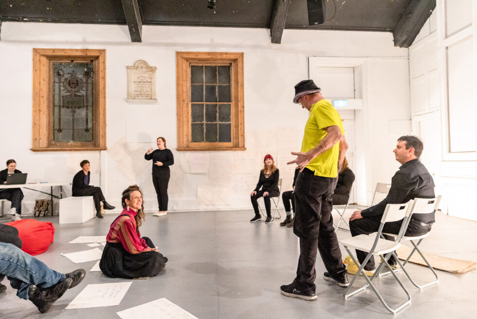 People standing and seated in an open hall rehearsing a performance.