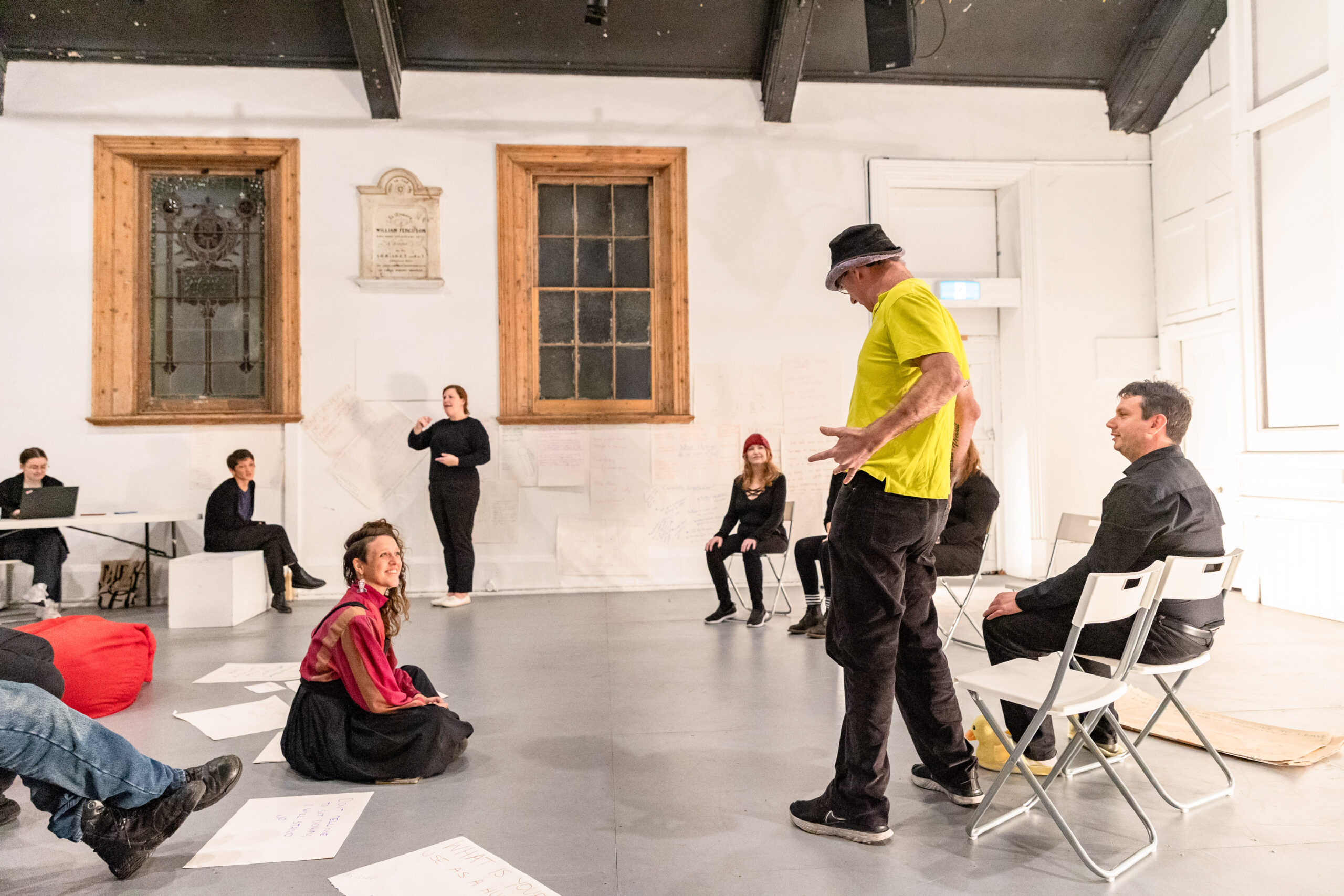 People standing and seated in an open hall rehearsing a performance.